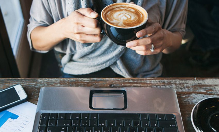 Eine Frau sitzt am Laptop und hält eine Tasse Kaffee in der Hand